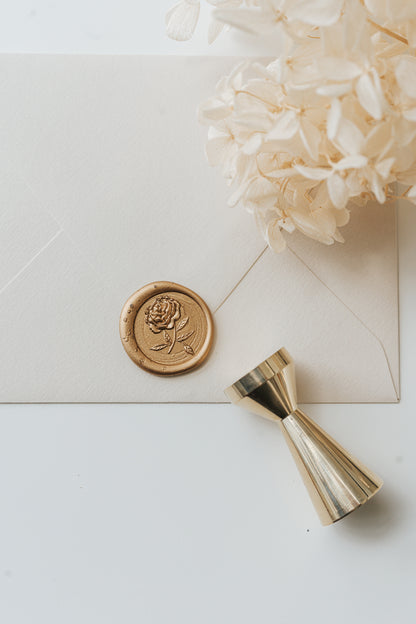 Gold peony wax seal on cream envelope beside brass wax seal stamp and dried hydrangea flowers