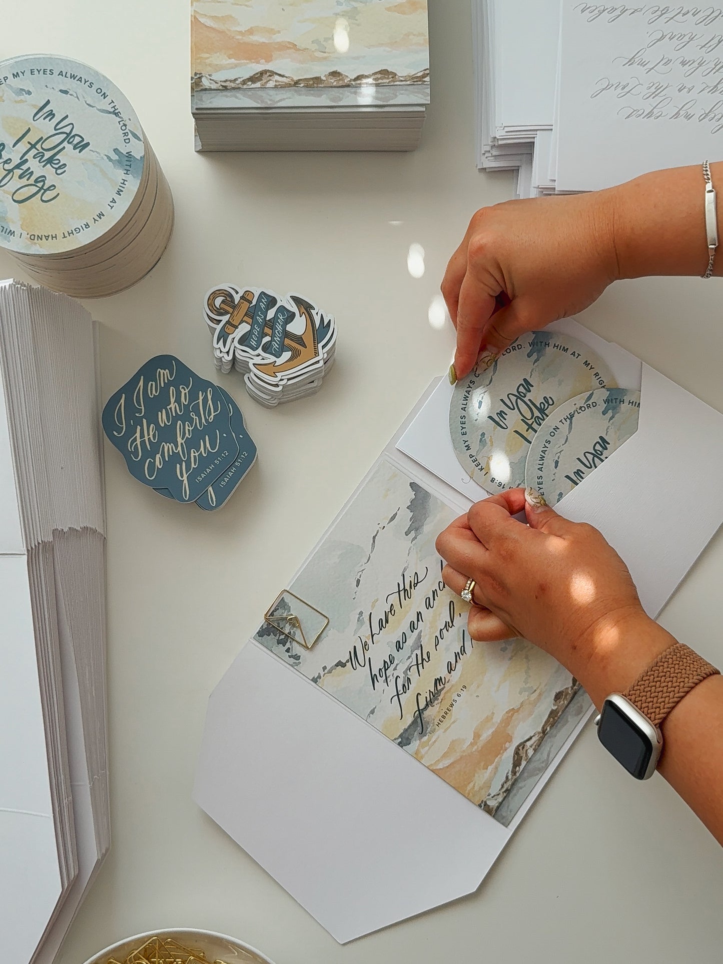 Hands assembling September Scribe snail mail kit with watercolor Hebrews 6:19 card, In You I Take Refuge coasters, Isaiah 51:12 sticker and Hope as an Anchor sticker