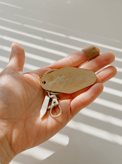 Personalized metal motel keychain with custom calligraphy name engraving, gold key ring, and calligraphy nib charm, shown in hand for size reference and everyday use.