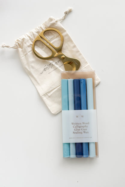 Gold brass wax cutter with a cotton storage pouch beside blue gradient glue gun sealing wax sticks for creating custom wax seals on wedding invitations, envelopes, and stationery.