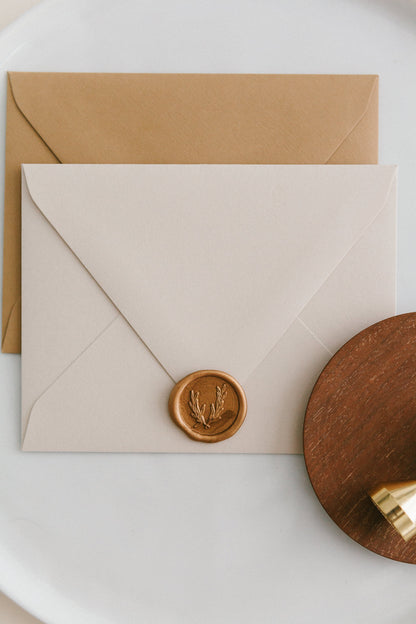 Brass wax seal stamp on a wooden dish beside envelopes with a gold olive crest wax seal, styled on a white ceramic plate.