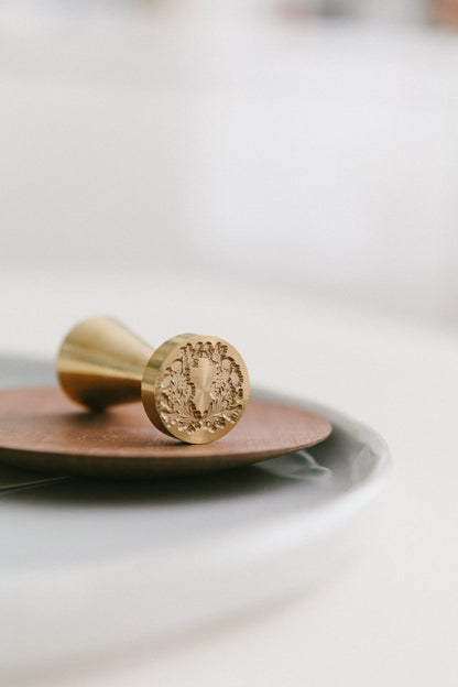 A brass wax seal stamp with floral crest design styled on a wooden and white ceramic plate