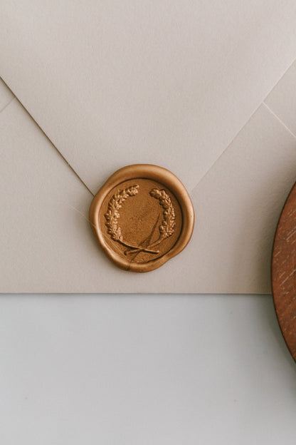 A close-up of a golden laurel crest wax seal, styled on a beige sealed envelope on a white surface