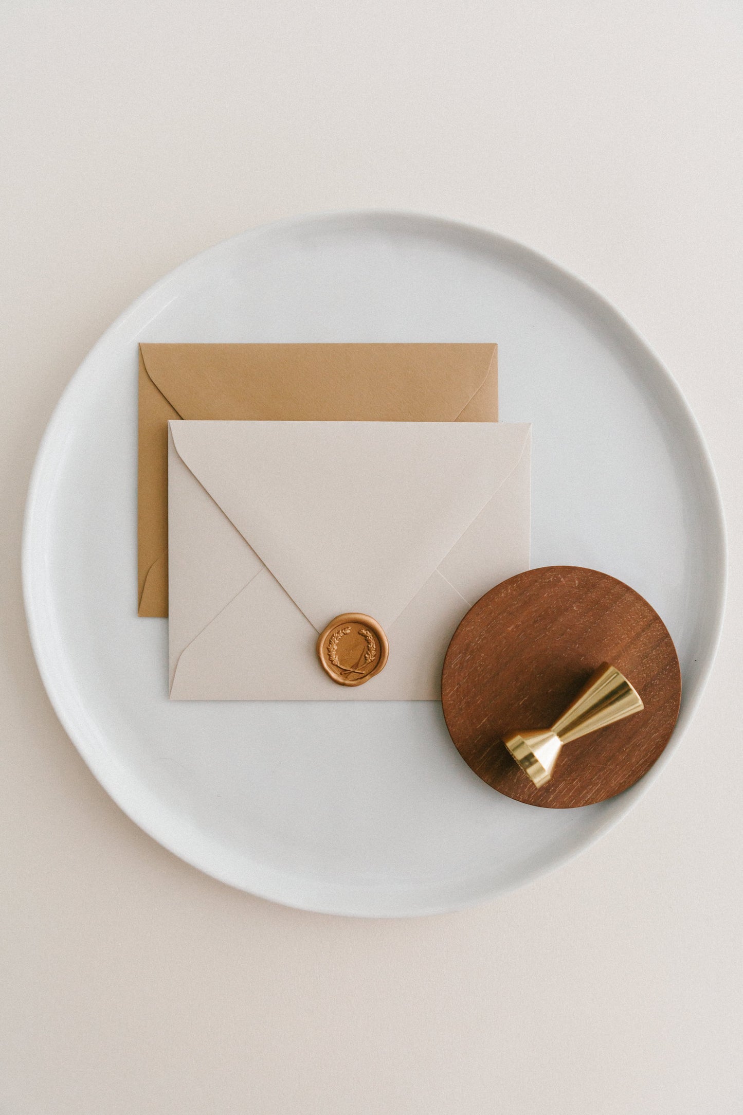 Brass wax seal stamp on a wooden dish beside envelopes with a gold laurel crest wax seal, styled on a white ceramic plate.
