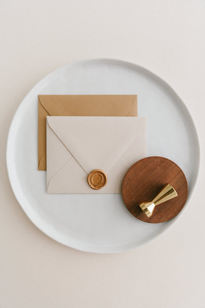 Brass wax seal stamp on a wooden dish beside envelopes with a gold laurel crest wax seal, styled on a white ceramic plate.