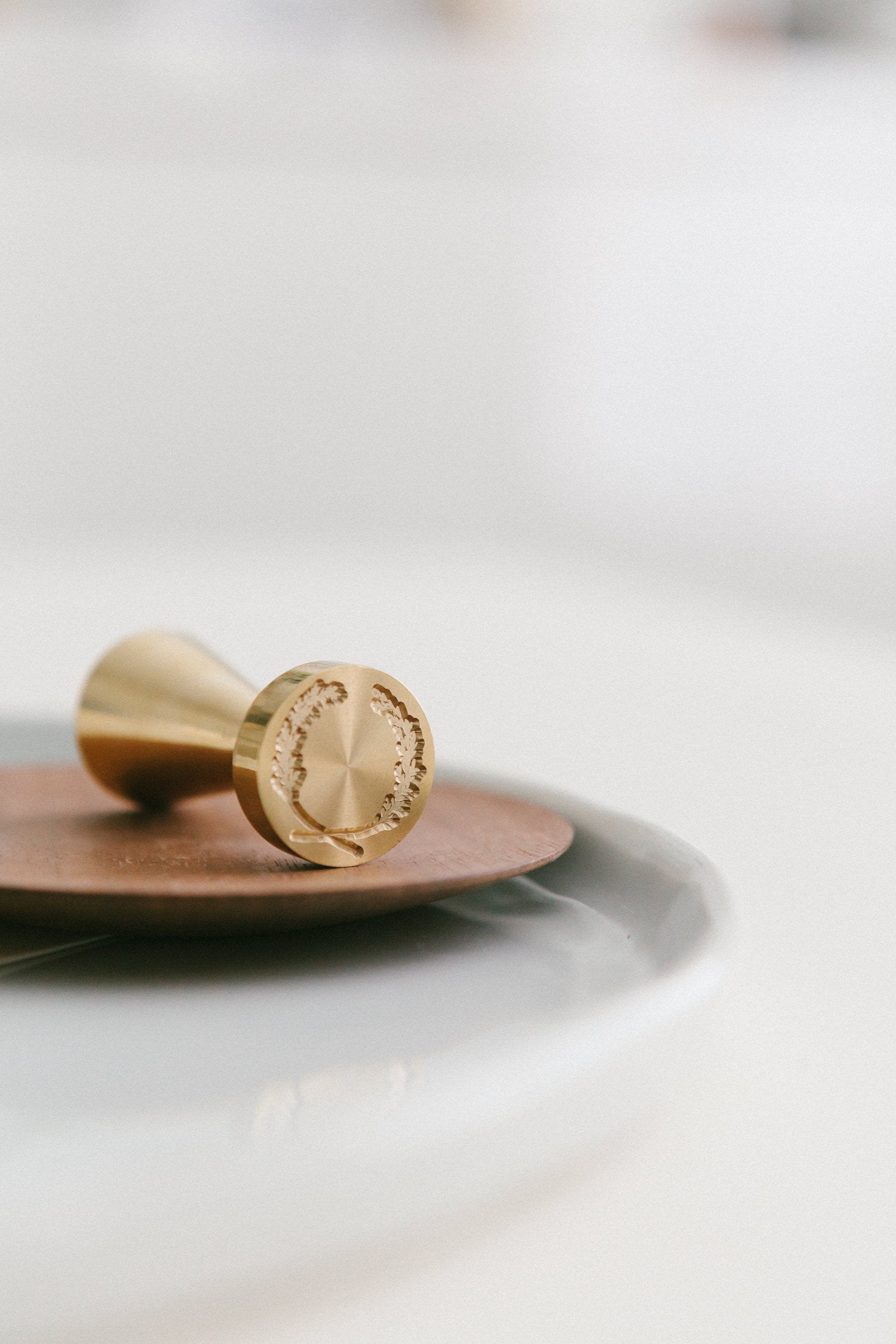 A brass wax seal stamp with laurel crest design styled on a wooden and white ceramic plate