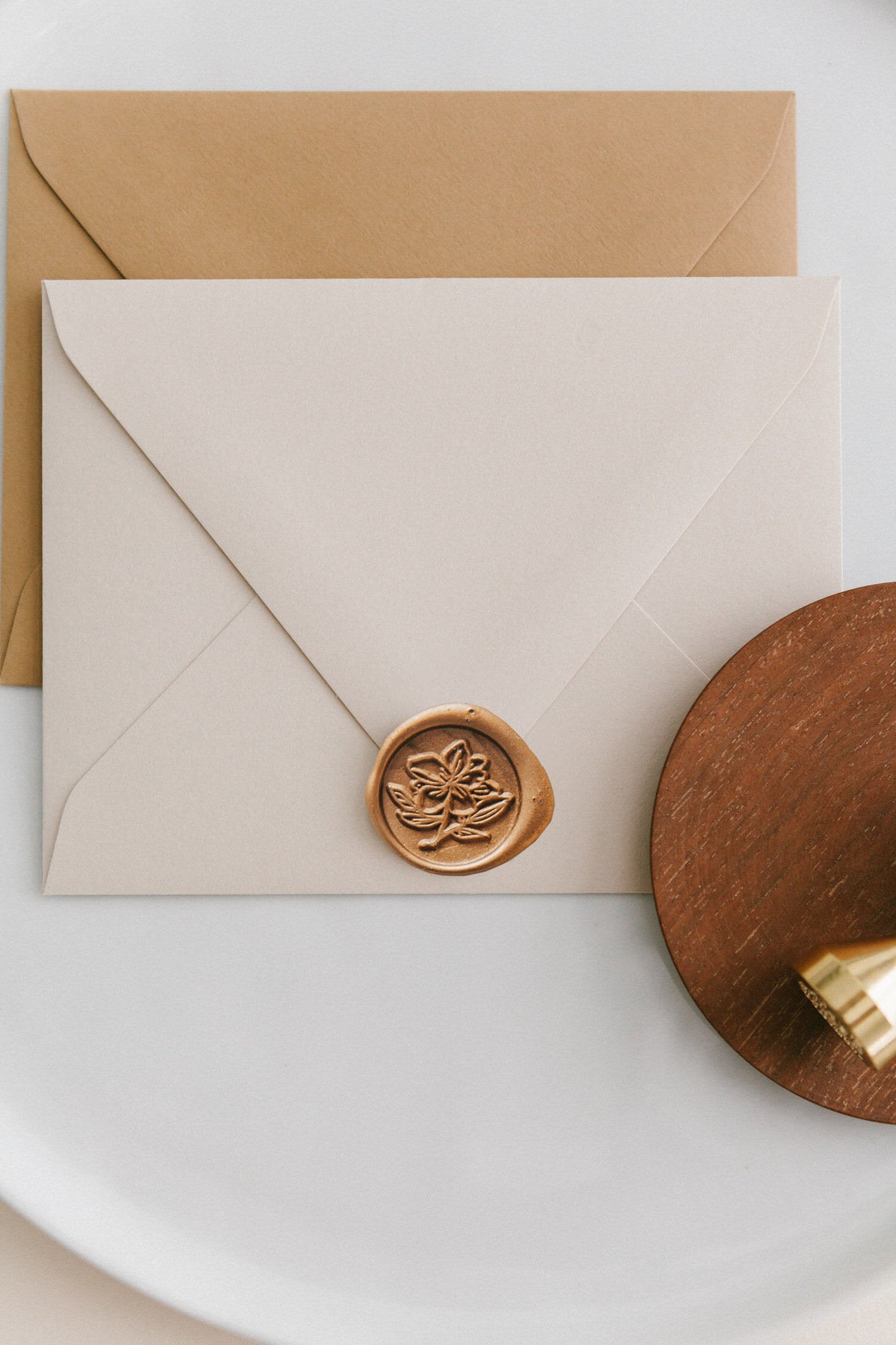 Brass wax seal stamp on a wooden dish beside envelopes with a gold magnolia wax seal, styled on a white ceramic plate.