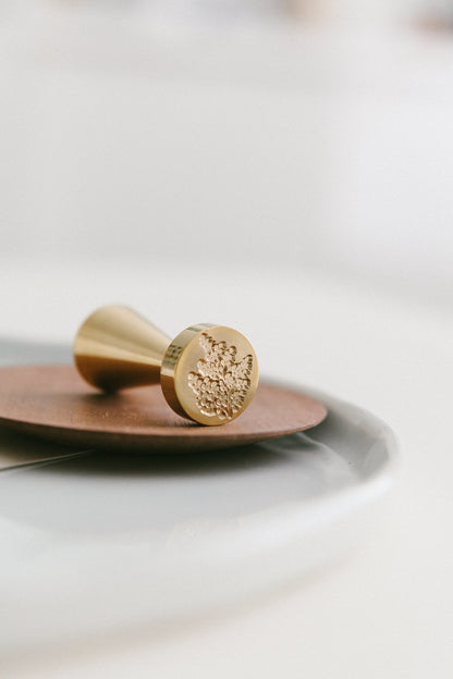 A close-up of a golden maidenhair fern wax seal stamp, styled on a wooden and white ceramic plate