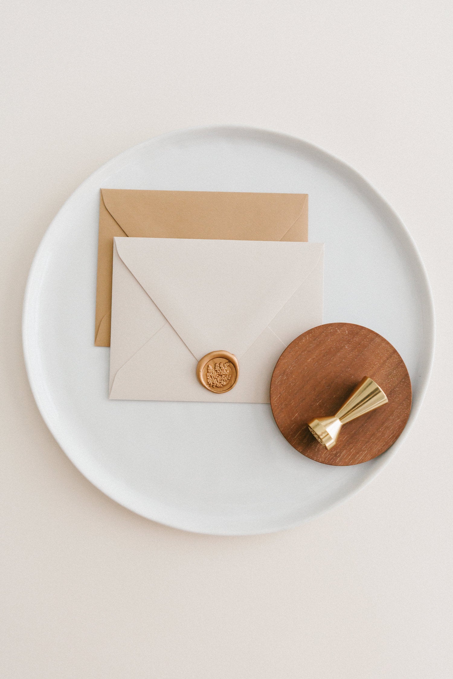 Brass wax seal stamp on a wooden dish beside envelopes with a gold maidenhair fern wax seal, styled on a white ceramic plate.