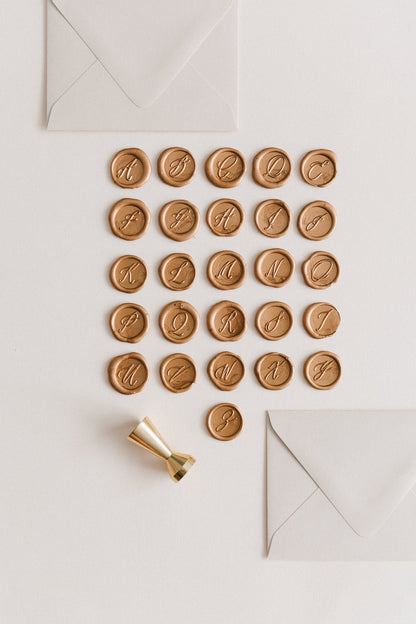 Gold monogram wax seals arranged in alphabetical order with a gold wax seal stamp and two ivory envelopes on a neutral background.