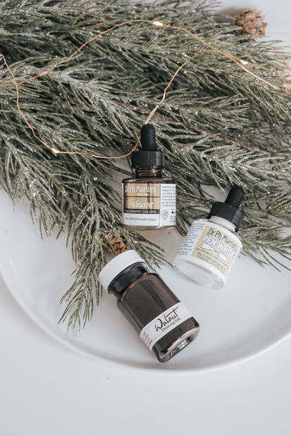 Dr. Ph. Martin's Iridescent, Walnut Drawing Ink, and Pen-White bottles with festive greenery and holiday decor