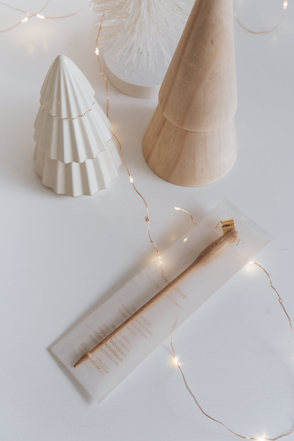 A wooden calligraphy pen in a clear package, surrounded by ceramic and wooden trees, with delicate string lights on a white surface