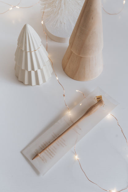 A wooden calligraphy pen in a clear package, surrounded by ceramic and wooden trees, with delicate string lights on a white surface