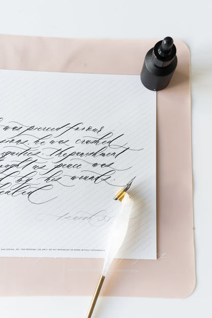 calligraphy pen rests on a slanted grid notepad with ink bottle and a pink silicone mat with 'practice makes progress' text