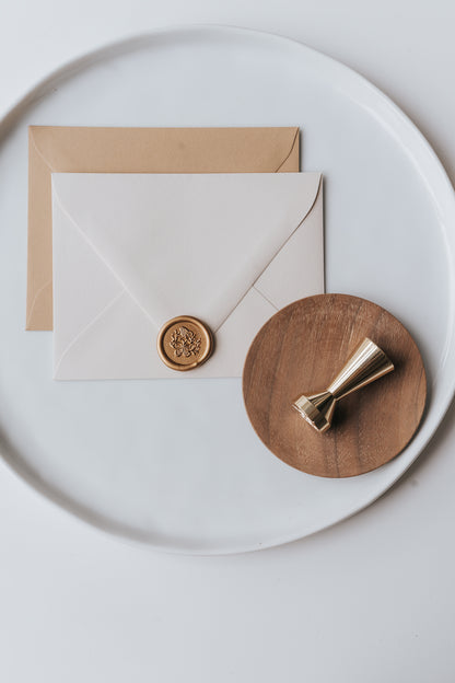 Brass wax seal stamp on a wooden dish beside envelopes with a gold butterflies wax seal, styled on a white ceramic plate.