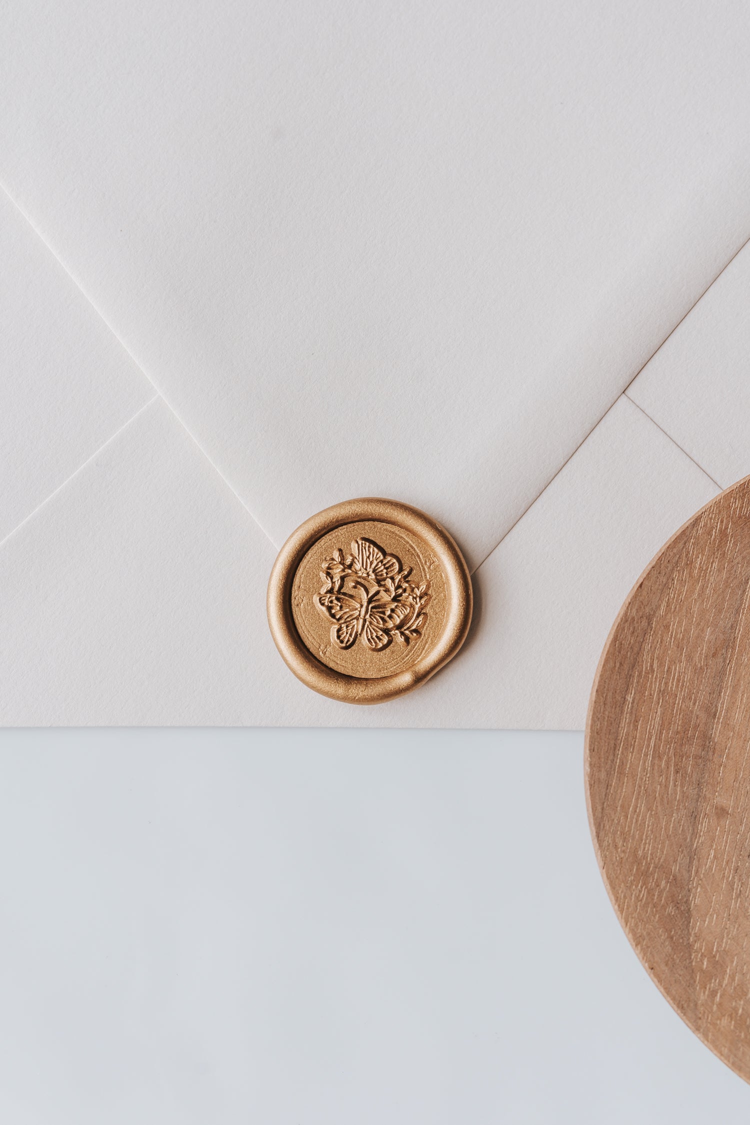 A golden butterflies wax seal, styled on a beige sealed envelope and a wooden dish on the side