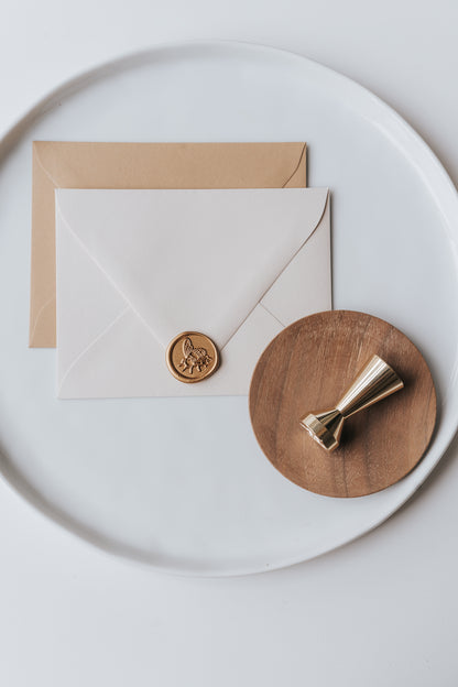 Brass wax seal stamp on a wooden dish beside envelopes with a gold honeybee wax seal, styled on a white ceramic plate.