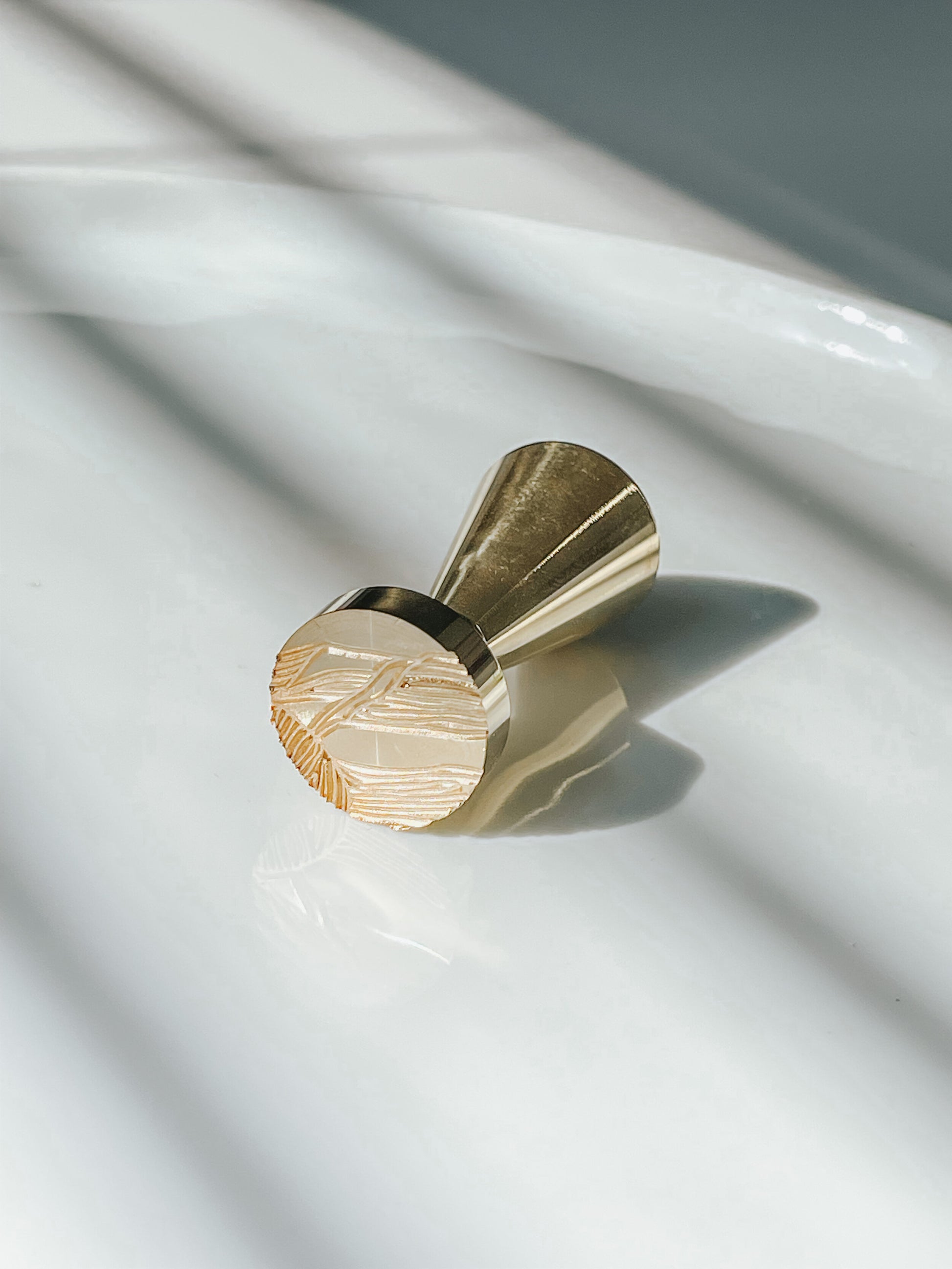 Gold brass sand dunes wax seal stamp resting on a glossy white surface, with sunlight casting soft shadows