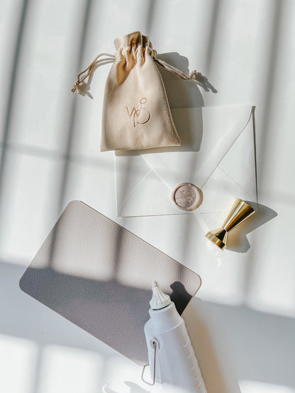 Wax sealing setup with a gold brass sand dunes wax stamp, envelope, suede pouch, and a white wax glue gun on a sunlit surface