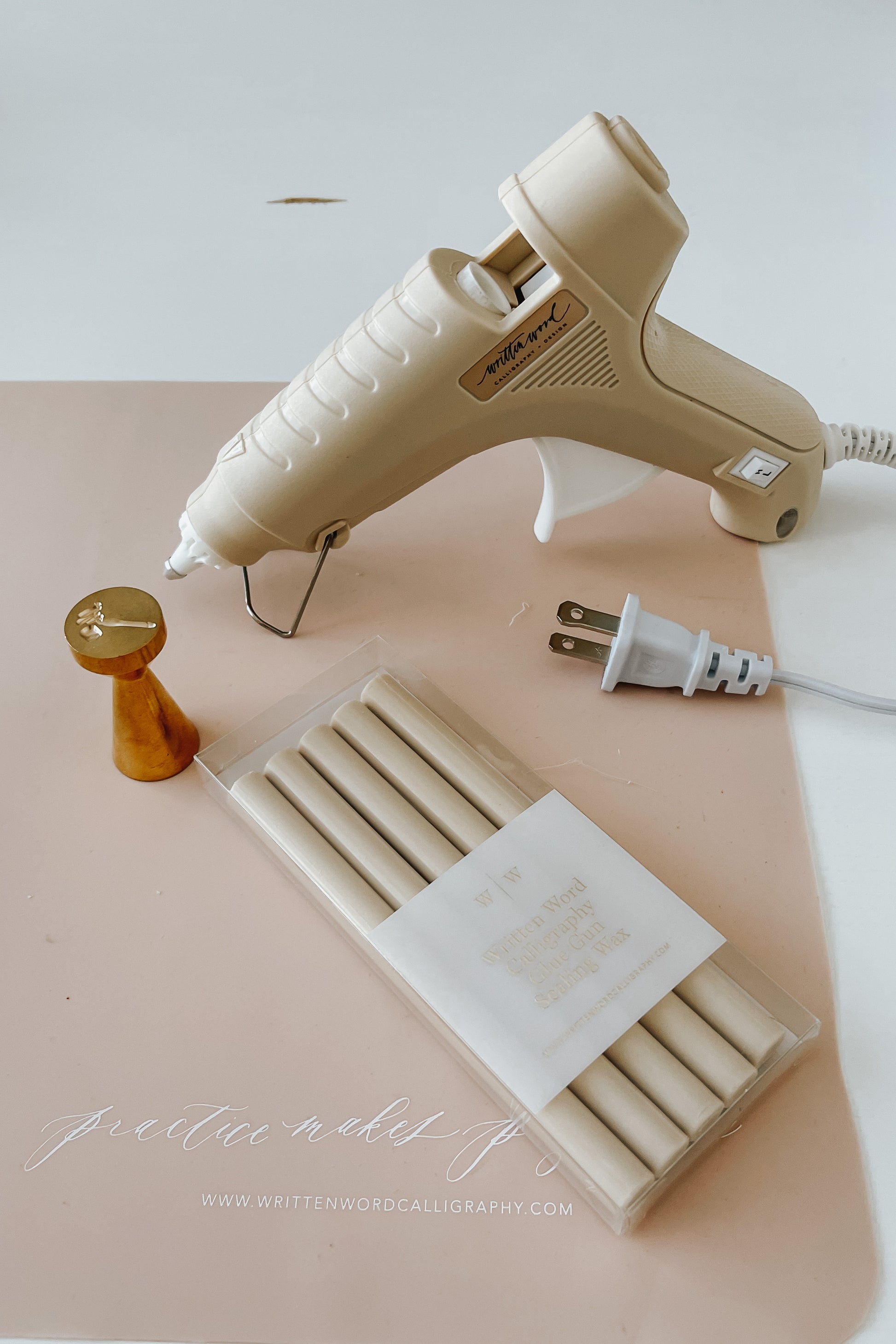 A beige Dual-Heat Glue Gun, a brass wax seal stamp and a pack of wax sealing sticks on a pink Written Word branded silicon mat