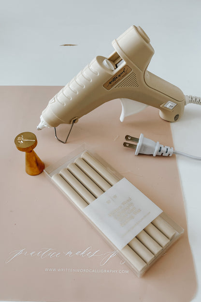 A beige Dual-Heat Glue Gun, a brass wax seal stamp and a pack of wax sealing sticks on a pink Written Word branded silicon mat