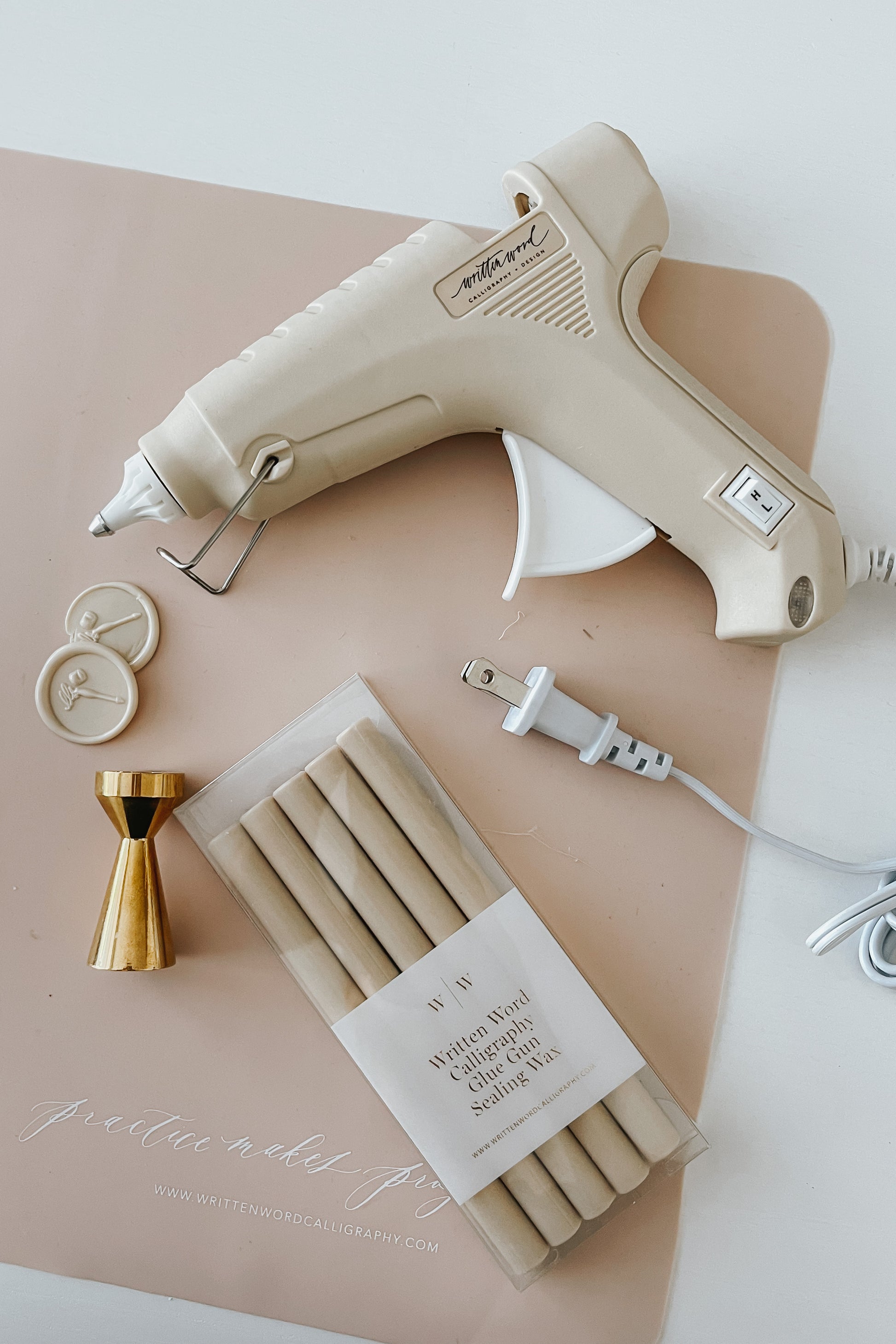 A beige Dual-Heat Glue Gun, wax seal stamps, beige wax seals and a pack of sealing wax sticks on top of a pink silicon mat