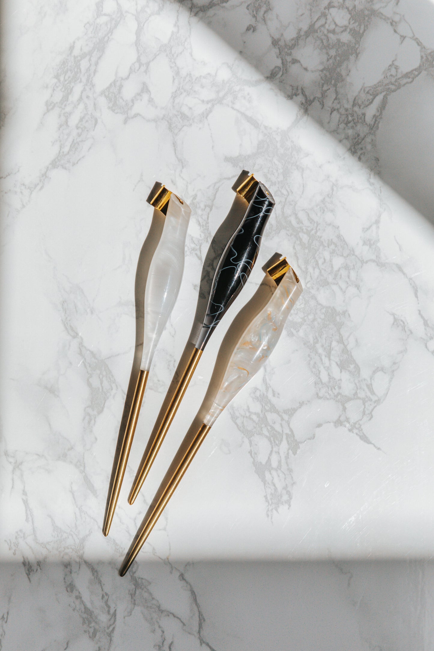 A trio of oblique calligraphy pens in white, black, and pearl tones with brass tips, showcased on a marble-patterned surface
