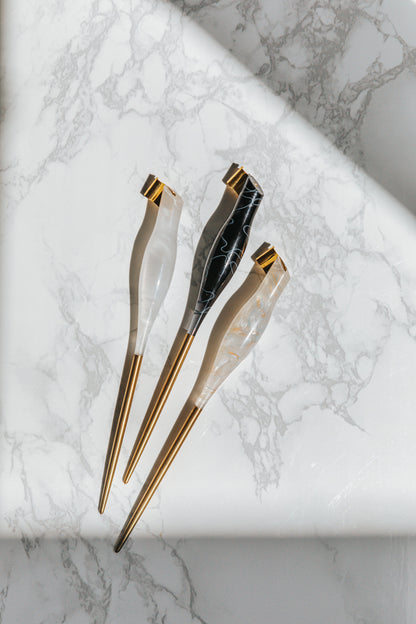 A trio of oblique calligraphy pens in white, black, and pearl tones with brass tips, showcased on a marble-patterned surface