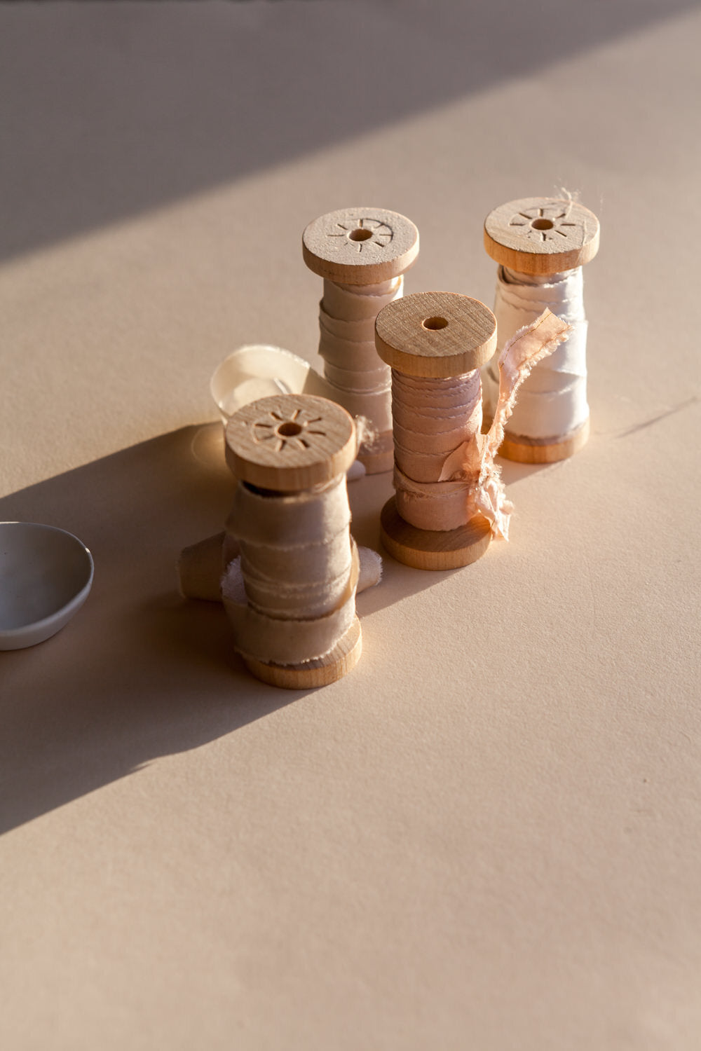 Wooden spools wrapped with silk ribbon in neutral tones, styled in warm sunlight on textured background