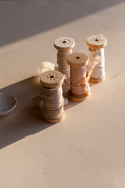 Wooden spools wrapped with silk ribbon in neutral tones, styled in warm sunlight on textured background