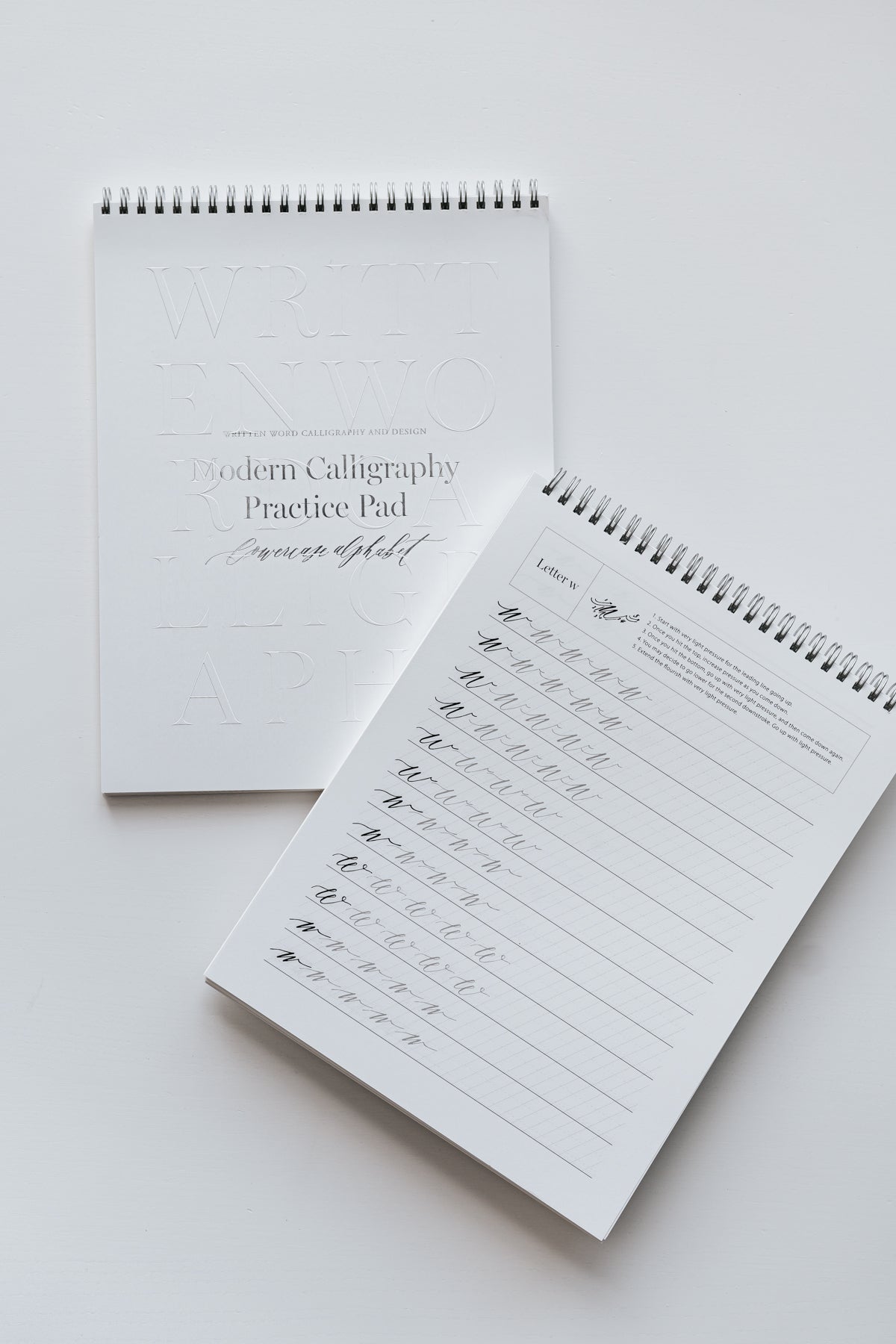 Two spiral-bound modern calligraphy practice pads, one closed and one open to a lowercase 'w' practice page on a white surface