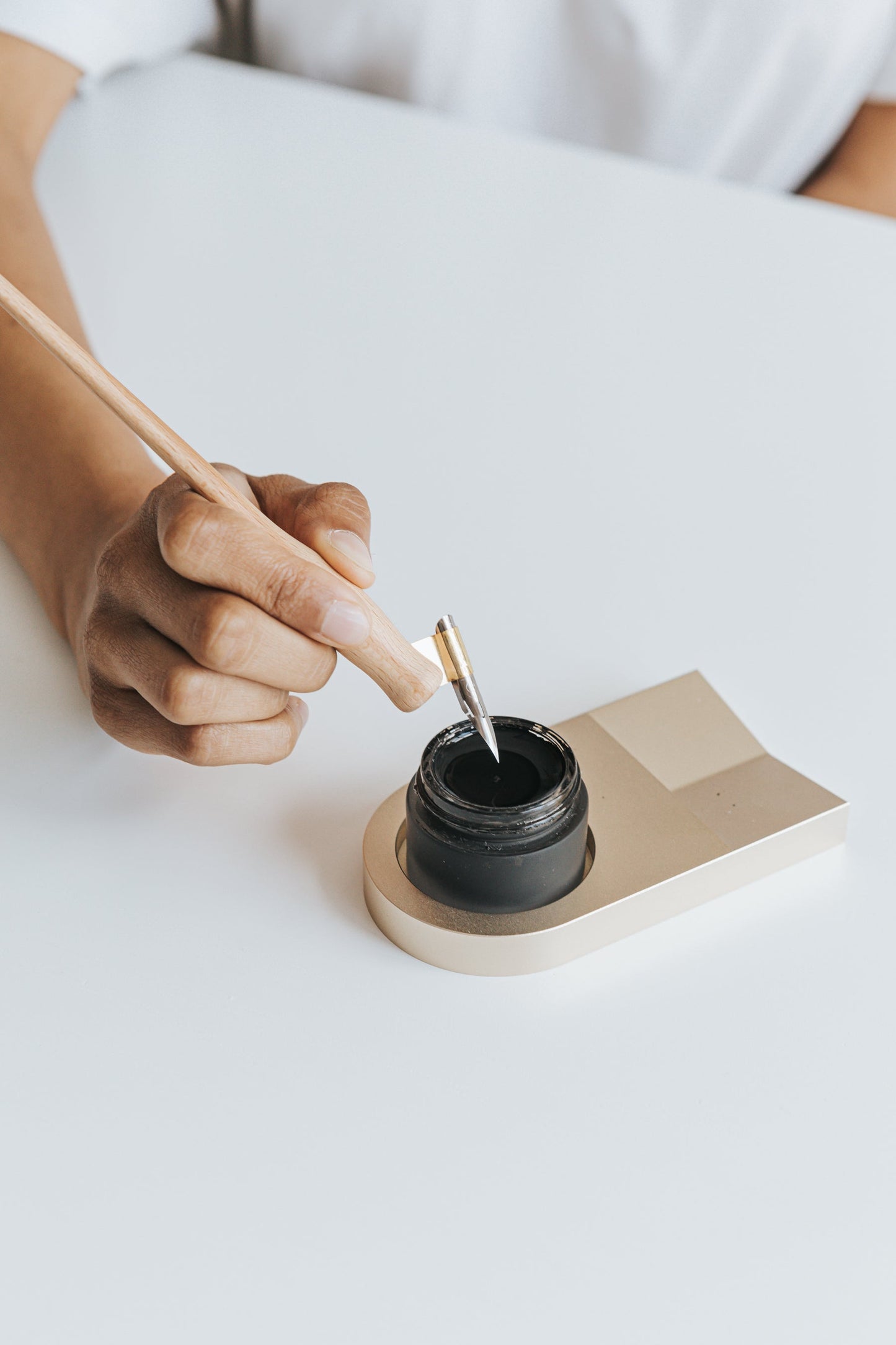 Hand holding a wooden calligraphy pen with a brass flange, dipping into an ink jar resting on a sleek gold inkwell holder