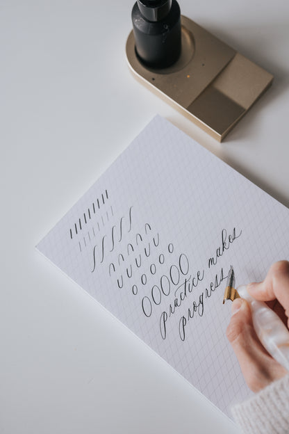 Hand practicing calligraphy on a slanted guide notepad, writing 'practice makes progress' with a pointed pen