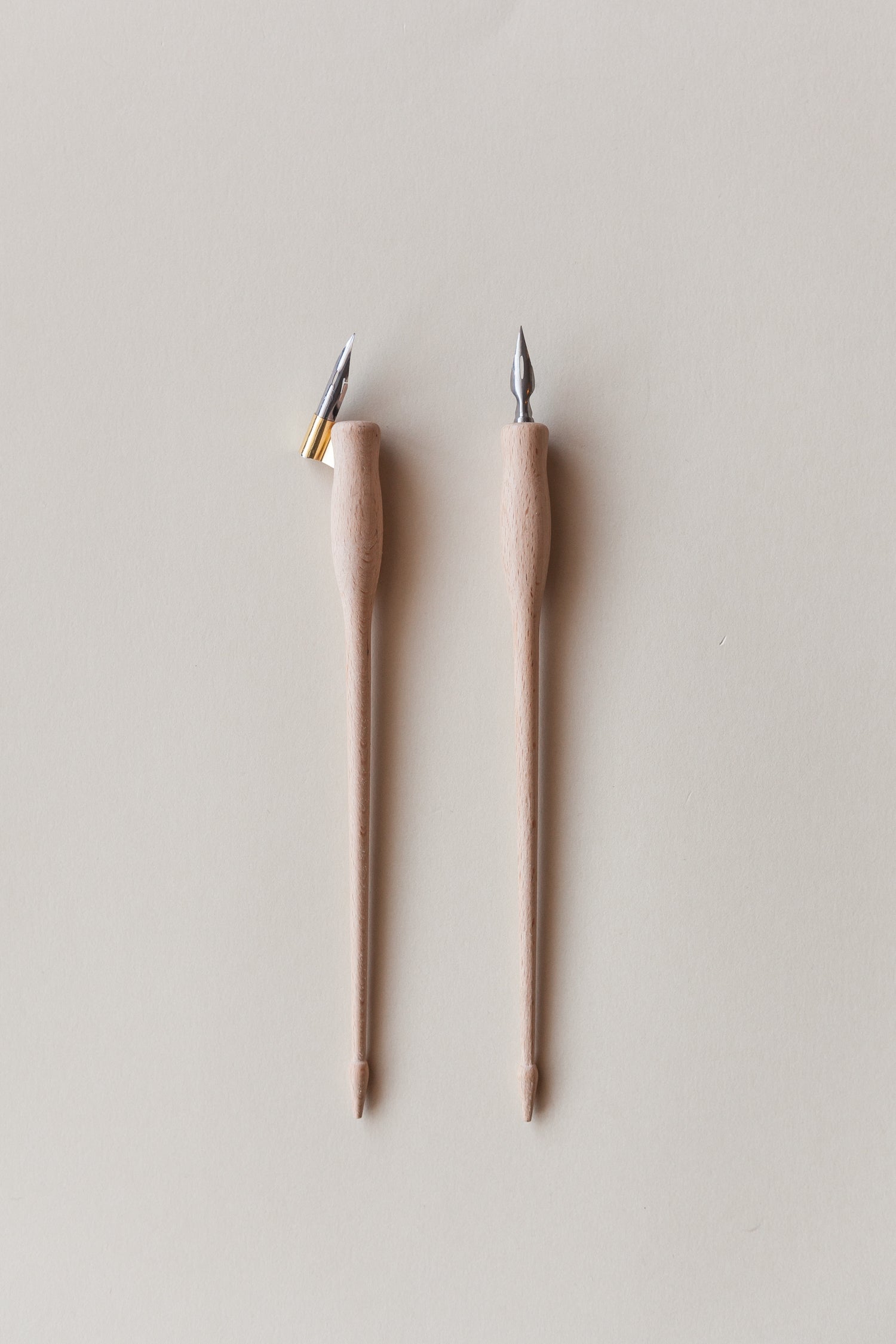 Two wooden calligraphy pens, one with a brass flange and the other straight, featuring metal nibs, on a neutral beige background