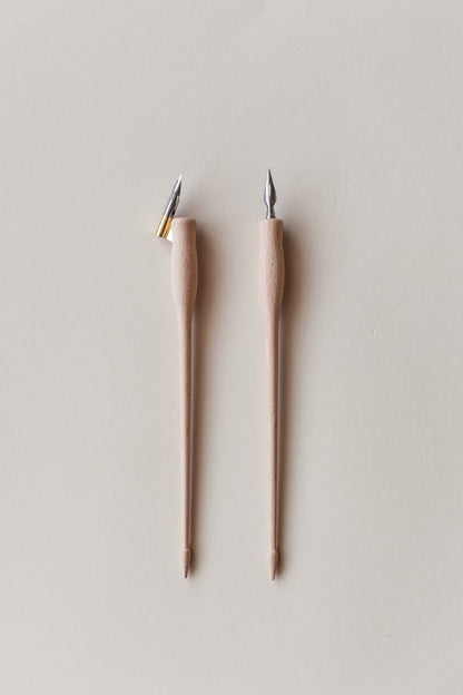 Two wooden calligraphy pens, one with a brass flange and the other straight, featuring metal nibs, on a neutral beige background
