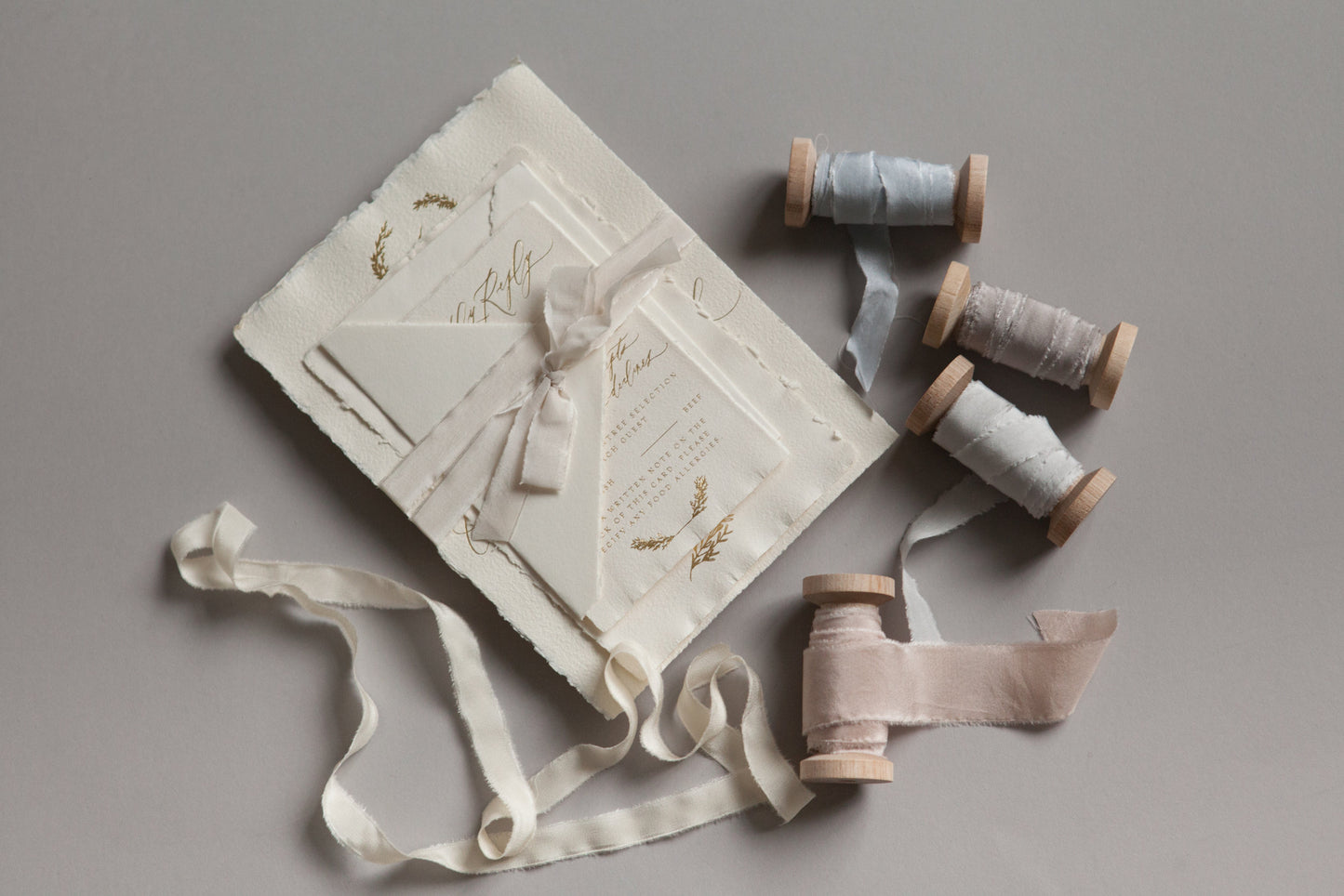 Handmade wedding invitation suite with deckled paper, silk ribbon, and wooden spools on soft neutral surface