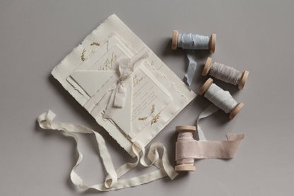 Handmade wedding invitation suite with deckled paper, silk ribbon, and wooden spools on soft neutral surface